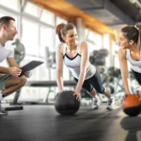 Two sporty women exercising with fitness balls while their fitness instructor tracking the progress on clipboard.